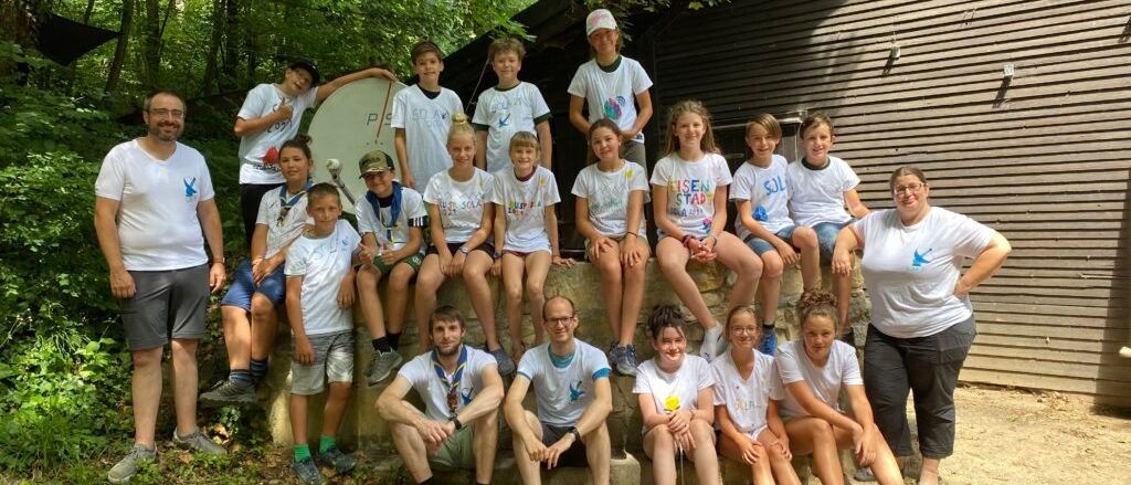 Eine Gruppe von Kindern und Erwachsenen posiert für ein Foto im Wald, alle tragen weiße T-Shirts. Sie sitzen auf einer Steinmauer.