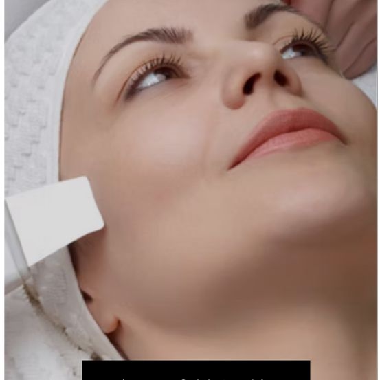 Eine Frau erhält eine Gesichtsbehandlung mit einem weißen Gerät, ihr Gesicht ist sauber und sie hat ein Handtuch auf dem Kopf.