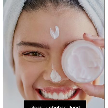 Eine Frau mit einem Handtuch auf dem Kopf lächelt und trägt eine weiße Creme auf ihr Gesicht auf.