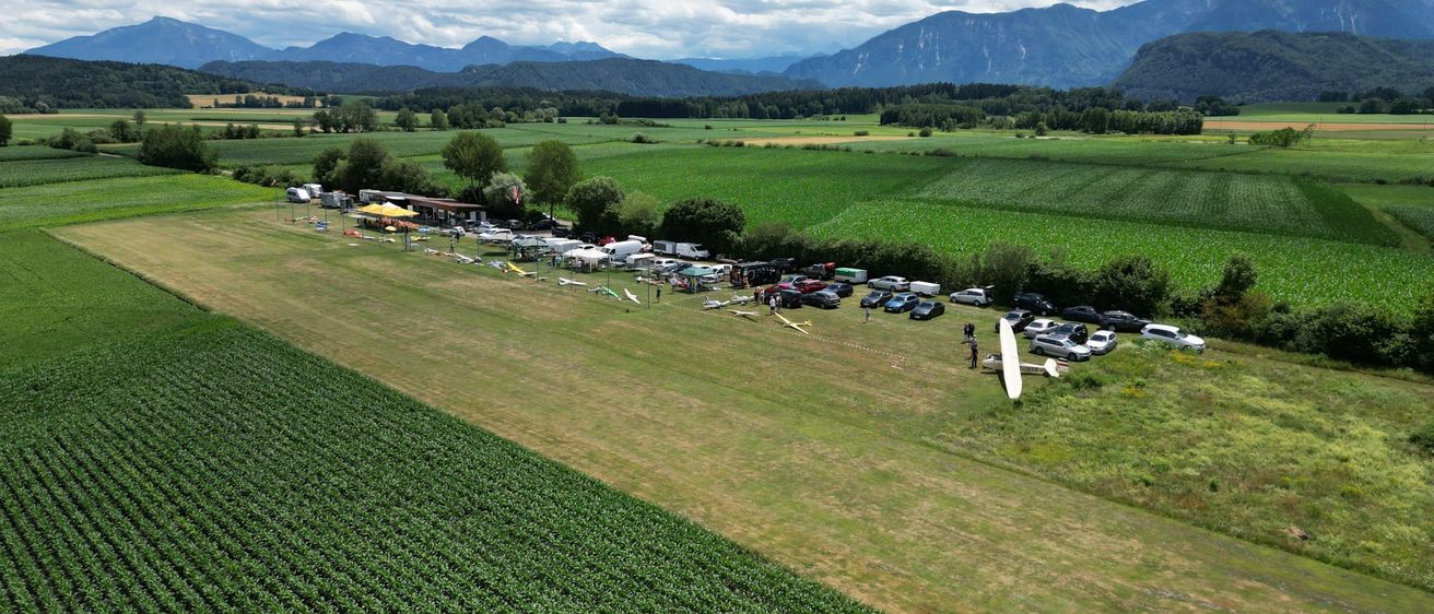 Luftaufnahme eines großen Feldes mit geparkten Autos, Zelten und Menschen. Ein Flugzeug steht auf dem Boden, mit Bergen im Hintergrund.