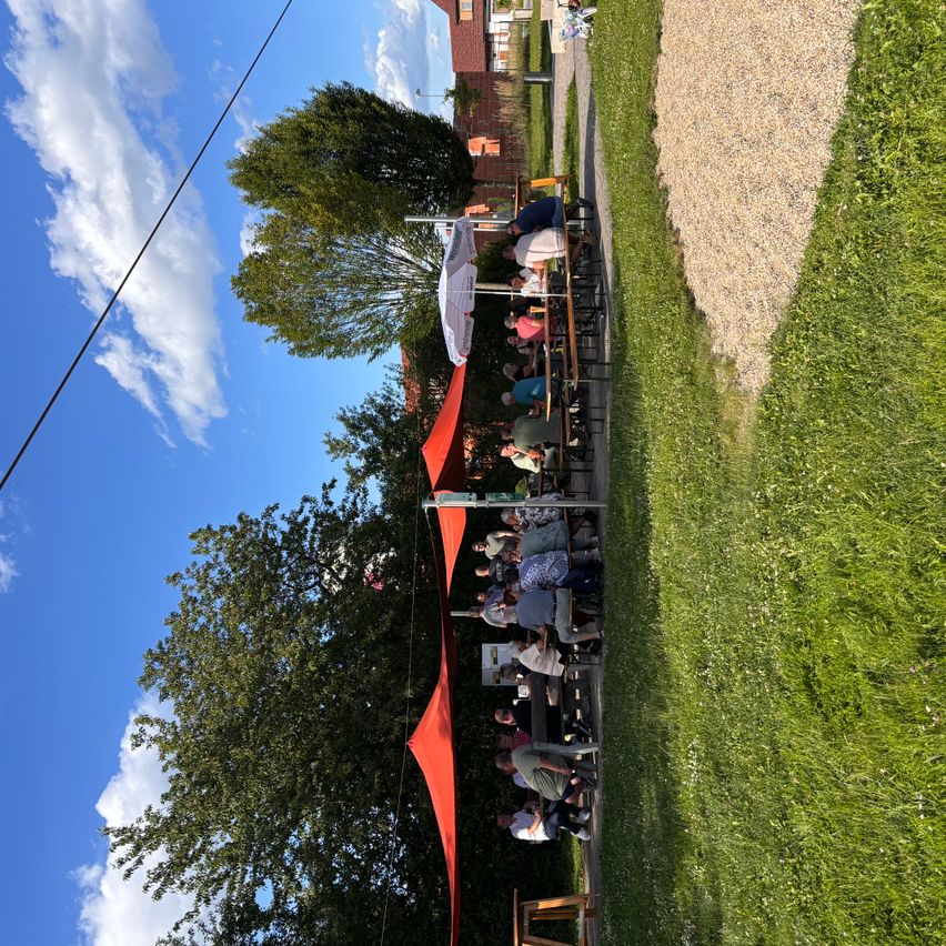 A group of people are sitting at tables under red canopies in a grassy area, with a building in the background. The sky is blue with some clouds.