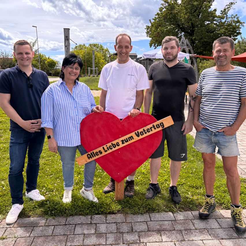 Five people stand on a brick pathway, smiling for a photo. They hold a red heart-shaped sign with German text. The setting is outdoors with green grass and trees in the background.