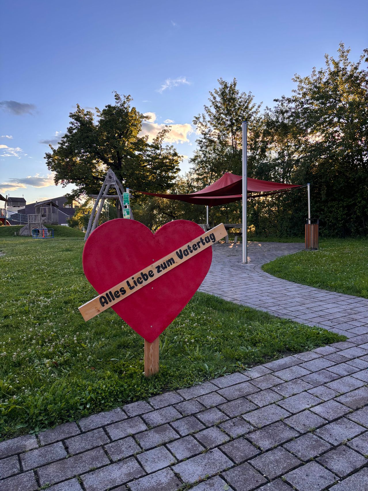 A red heart-shaped sign on a grassy field with a wooden post. Behind it is a pathway with a red canopy, trees, and buildings. The sign reads 'Alles Liebe zum Vatertag', a German phrase meaning 'All Love to Father's Day'.