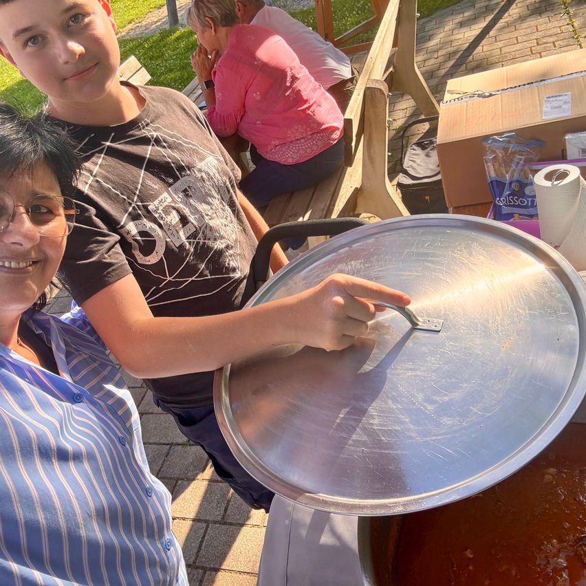 A boy and a woman are smiling in front of a large pot with a lid. Behind them, another woman is sitting on a bench.