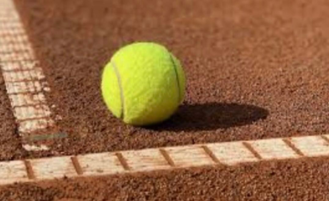 Ein Tennisball liegt auf einem Sandplatz, nahe einer Linienmarkierung. Der Ball ist gelb mit grünen Markierungen.