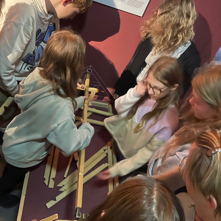 Kinder beschäftigen sich mit einer praktischen Aktivität, bei der sie ein Holzgerüst auf einem Tisch in einem Museum bauen. Sie beobachten und besprechen ihre Kreation genau.