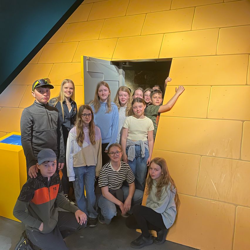 Eine Gruppe von Kindern und Jugendlichen posiert für ein Foto vor einer gelben Ziegelwand mit einer offenen Tür. Sie scheinen sich in einem Museum zu befinden.