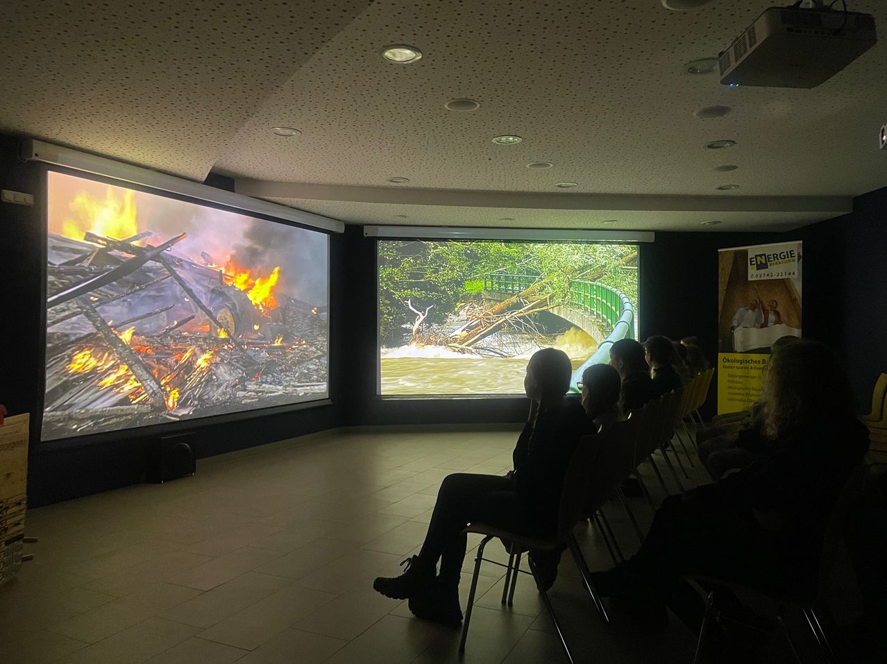 Ein Innenkino zeigt einen Dokumentarfilm, zwei große Bildschirme zeigen ein brennendes Auto und einen überfluteten Fluss, während die Leute auf Stühlen sitzen und der Präsentation zuschauen.