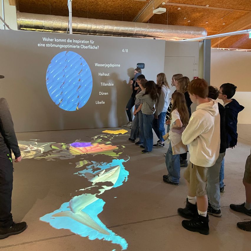 Eine Gruppe von Kindern beobachtet eine interaktive Anzeige einer Karte mit Wasserströmungen, die auf den Boden projiziert wird. Sie stehen vor einer Wand mit einer Frage zur Optimierung der Wasseroberfläche.