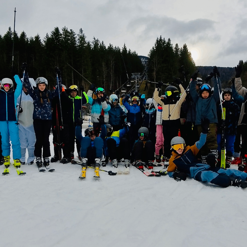 Eine Gruppe von Skifahrern, gekleidet in warme Winterkleidung und Helmen, posiert für ein Foto auf einem verschneiten Berg. Einige sitzen, andere stehen und halten Skistöcke. Im Hintergrund sind Kiefern und ein bewölkter Himmel zu sehen.