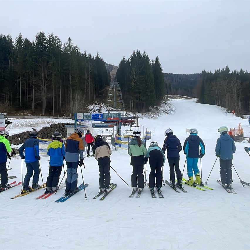 Eine Gruppe von Skifahrern wartet am Fuße einer Skipiste. Bäume säumen den Hintergrund, und eine Skiliftanlage ist in der Ferne zu sehen.