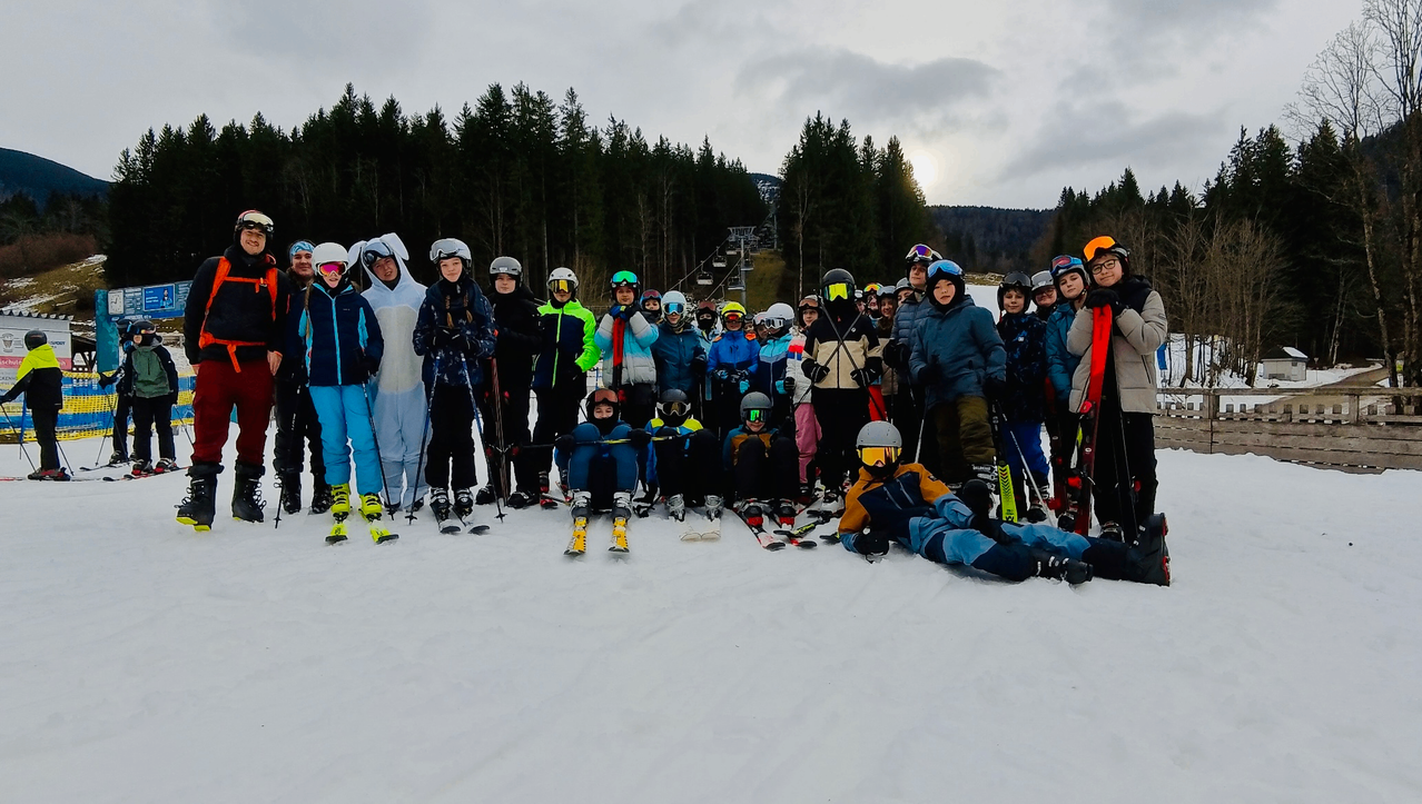 Eine Gruppe von Menschen in Skiausrüstung steht und sitzt auf einem verschneiten Hang mit Tannen im Hintergrund.
