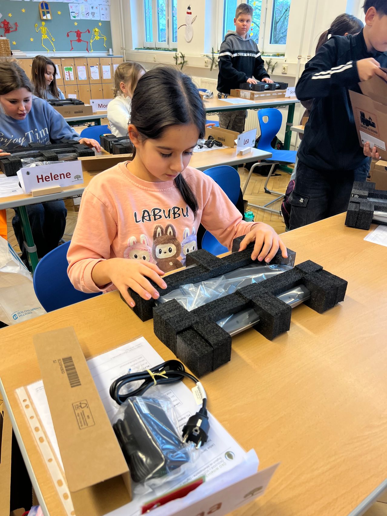 Ein junges Mädchen arbeitet an einem Laptop auf einem Schreibtisch. Andere Kinder sind im Hintergrund zu sehen, die ebenfalls an ähnlichen Aufgaben arbeiten.