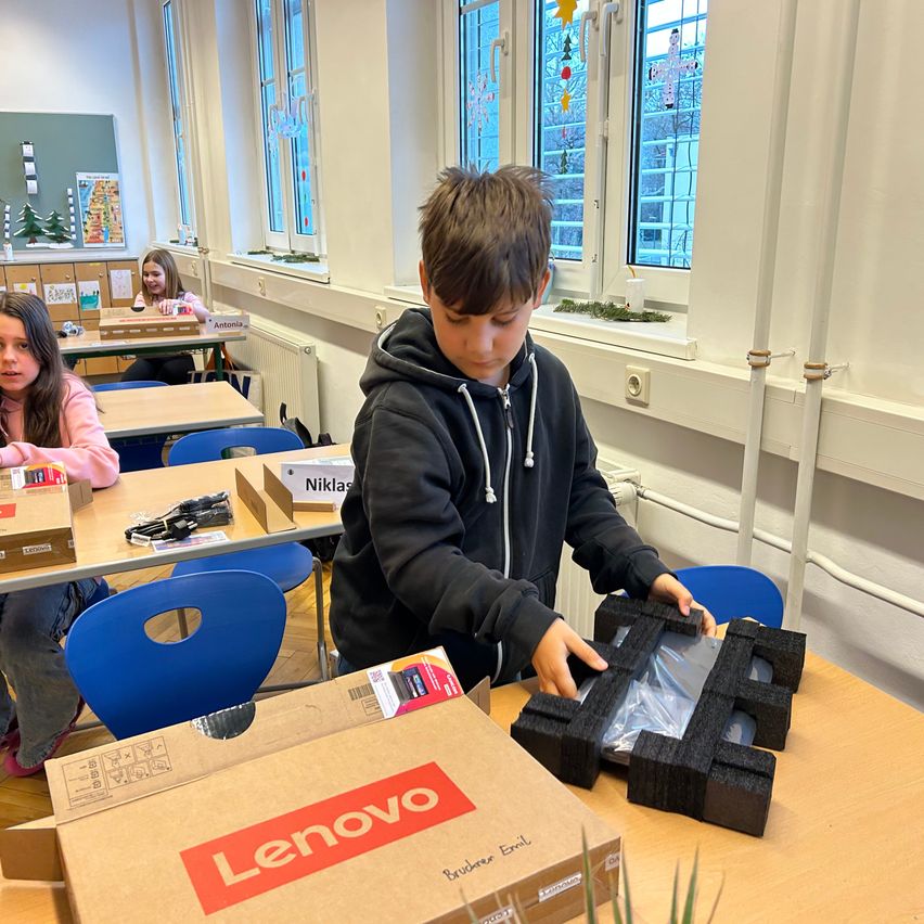Ein Junge in einem Klassenzimmer öffnet eine Lenovo-Laptop-Box, während er an einem Tisch sitzt. Zwei Mädchen sitzen hinter ihm an Schreibtischen.