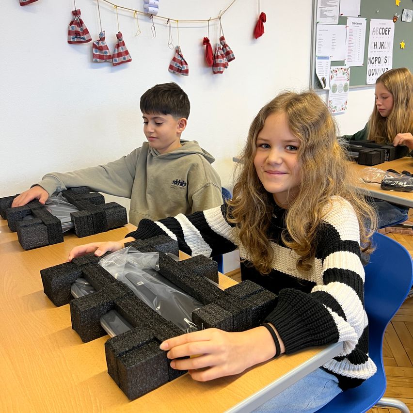 Ein junges Mädchen in einem schwarz-weißen Pullover lächelt, während sie und ein Junge ein Projekt mit Schaumstoffblöcken an einem Tisch bearbeiten. Hinter ihnen sitzt eine weitere Mädchen, und verschiedene Papiere sind an einer Pinnwand befestigt.