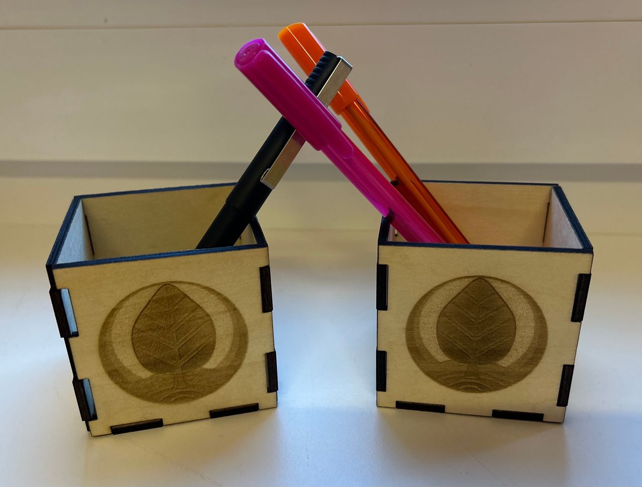 Zwei hölzerne Stifthalter mit Blattdesign auf einer weißen Oberfläche. Einer hält einen rosa und orange Stift, der andere einen schwarzen Stift.