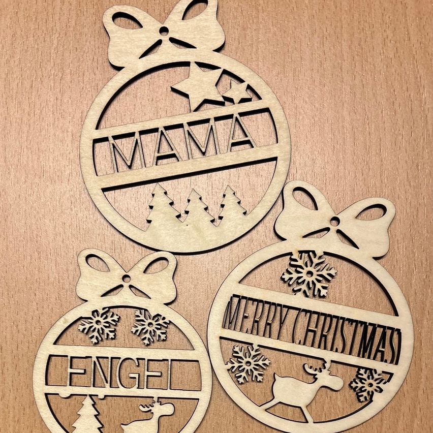 Drei Holzornamente auf einer Holzoberfläche. Eines steht 'MAMA', ein anderes 'ENGEL' und das letzte 'FROHE WEIHNACHTEN'. Jedes hat eine Schleife, einen Stern und Schneeflocken.