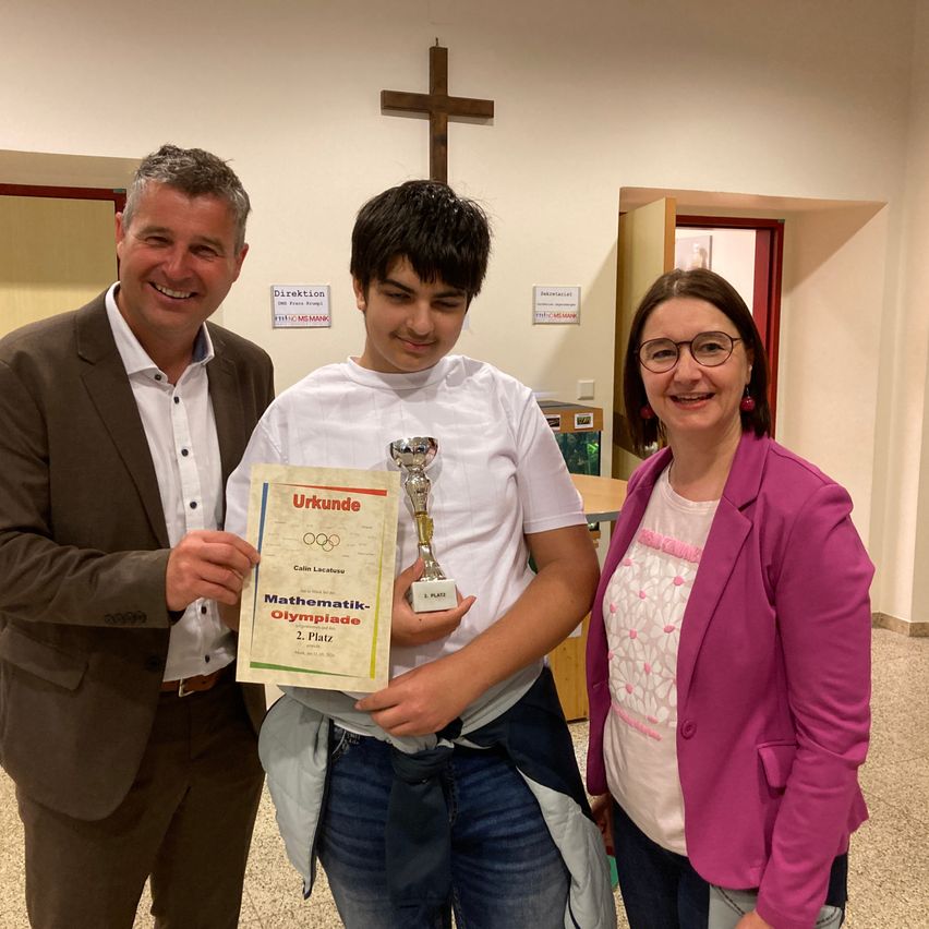 Ein junger Mann hält eine Trophäe und ein Zertifikat in der Hand und steht zwischen zwei lächelnden Erwachsenen in einem Raum mit einem Kreuz an der Wand.