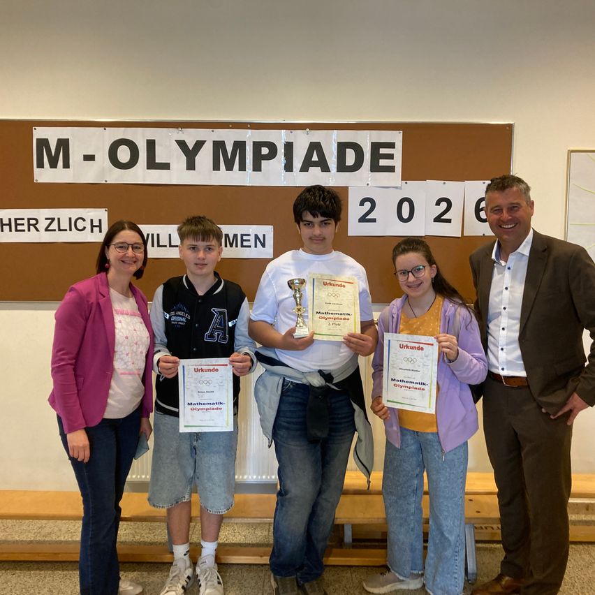 Fünf Personen stehen vor einem Schild, das 'M-OLYMPIADE 2026' besagt. Drei halten Urkunden, einer eine Trophäe, und der Mann lächelt. Sie tragen Freizeitkleidung.