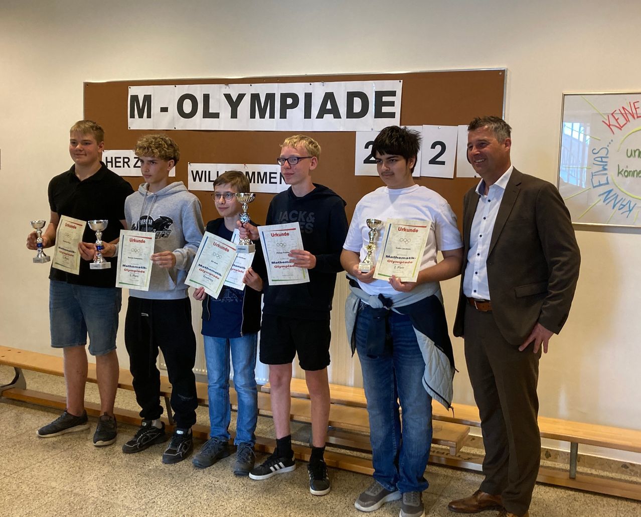 Sechs junge Männer stehen zusammen und halten Pokale und Urkunden. Sie stehen vor einem Schild mit den Worten 'M-Olympiade'. Sie tragen Freizeitkleidung und Schuhe. Hinter ihnen steht ein Mann im Anzug.