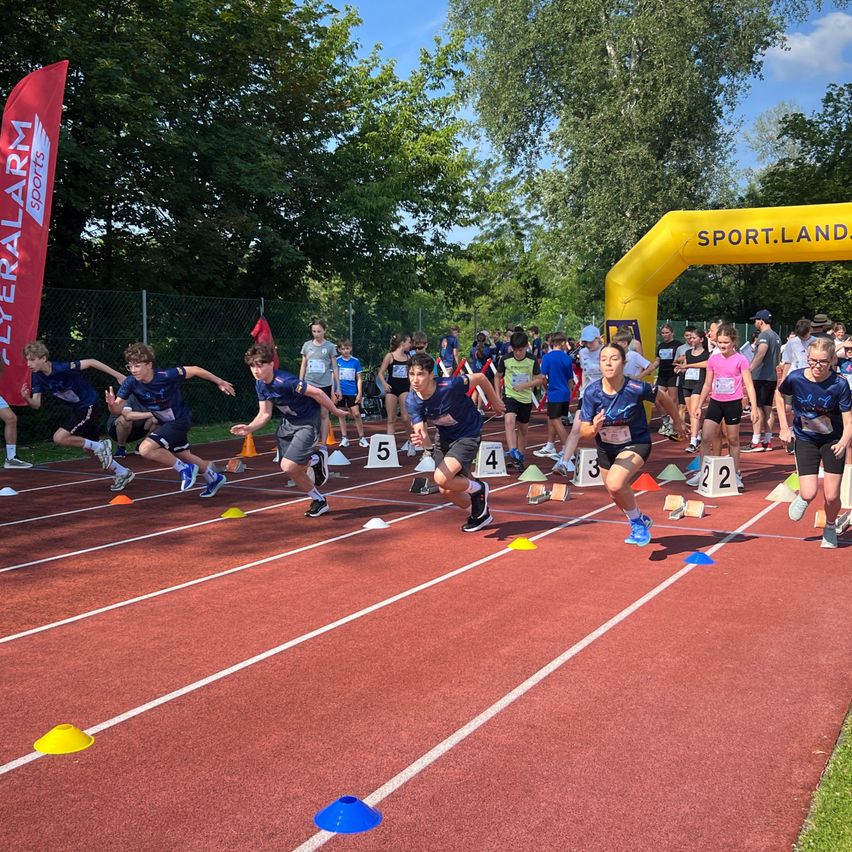 Eine Gruppe von Kindern läuft auf einer Rennbahn. Sie sind nummeriert von 1 bis 5. Sie tragen Turnschuhe und Startnummern. Hinter ihnen sind Bäume und ein Zaun. Ein gelber Bogen mit dem Text Sport.Land steht am Ende der Bahn.