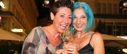 Zwei Frauen mit gefärbten Haaren, eine mit blauen und die andere mit blonden, lächeln und halten ein Weinglas, während sie für ein Foto posieren.