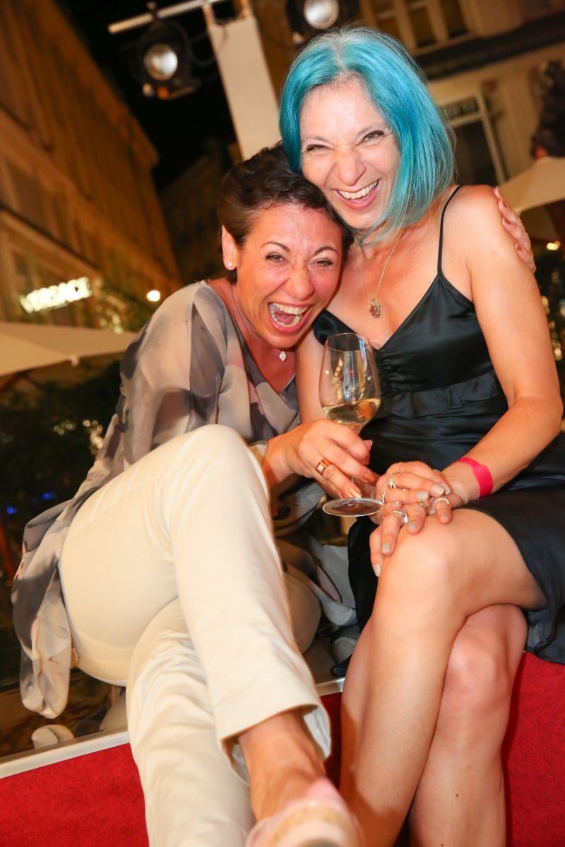 Two women laughing and posing for a photo, one holding a wine glass. They wear casual attire and appear to be friends enjoying an evening out.