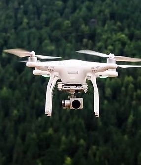 Ein weißer Drohne mit vier Propellern und einer Kamera fliegt über einen dichten Wald. Der Himmel ist klar.