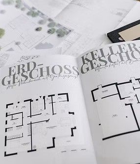 Geöffnetes Buch mit dem Titel 'Erdgeschoss' und Grundrissen eines Gebäudes, auf einem Tisch neben anderen Papieren ausgelegt.