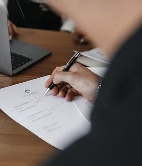Eine Person unterschreibt ein Dokument auf einem Schreibtisch mit einem Laptop und Papieren. Sie trägt einen schwarzen Anzug und hält einen Stift. Das Dokument enthält Text und ein Logo.