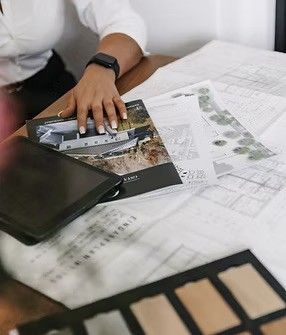 Eine Person sitzt an einem Schreibtisch mit Architekturplänen, einem Tablet und einer Farbpalette. Die Person trägt eine Uhr und einen Ring.