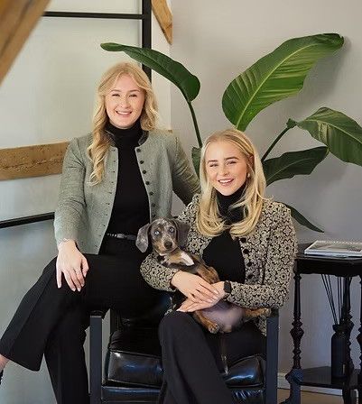 Zwei Frauen sitzen auf einem Stuhl und lächeln für ein Foto. Eine hält einen kleinen Hund. Sie tragen schwarze Outfits, eine in einer grünen Jacke. Eine große Pflanze und ein Holztisch stehen hinter ihnen.