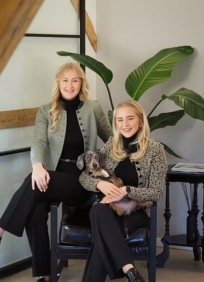 Zwei Frauen sitzen auf einem Stuhl und lächeln für ein Foto. Eine hält einen kleinen Hund. Sie tragen schwarze Outfits, eine in einer grünen Jacke. Eine große Pflanze und ein Holztisch stehen hinter ihnen.