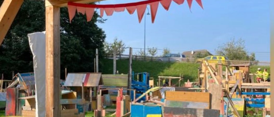 Ein Blick von innen aus einer Holzstruktur zeigt einen Rasenbereich mit verstreuten Holzkonstruktionen und Tischen. Rote dekorative Flaggen hängen von der Decke. In der Ferne sind eine Straße und geparkte Autos zu sehen.