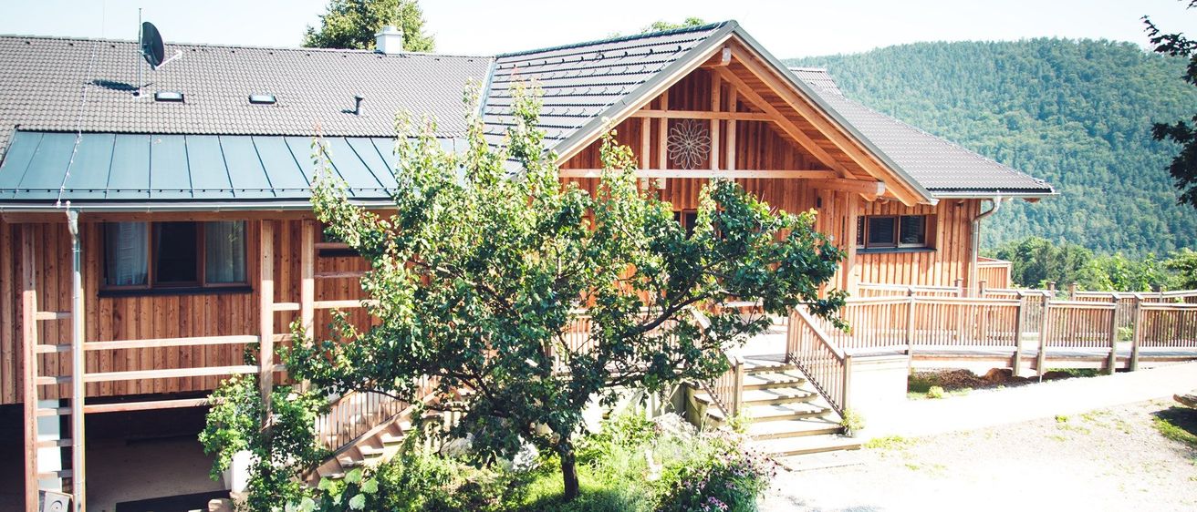Ein Holzhaus mit einem grauen Dach und einem gut gepflegten Garten davor. Das Haus hat einen Holzzaun und eine Treppe, die zum Eingang führt.