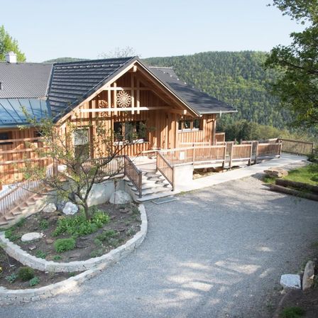 Ein Holzchalet mit grauem Dach und einem Blumendesign an der Vorderwand, umgeben von einem Garten und einer Schotterauffahrt, mit einem Berg im Hintergrund.