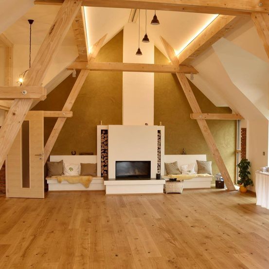 Ein leeres Dachzimmer mit Holzbalken, einem Kamin und zwei weißen Sofas mit Kissen. Der Boden ist aus Holz, und rechts steht eine Topfpflanze.