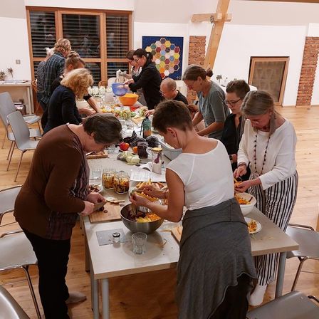 Eine Gruppe von Menschen versammelt sich um einen langen Tisch in einem Raum und bereitet Essen zu. Sie tragen Schürzen und scheinen an einer Kochaktivität beteiligt zu sein. Verschiedene Zutaten und Utensilien befinden sich auf dem Tisch.