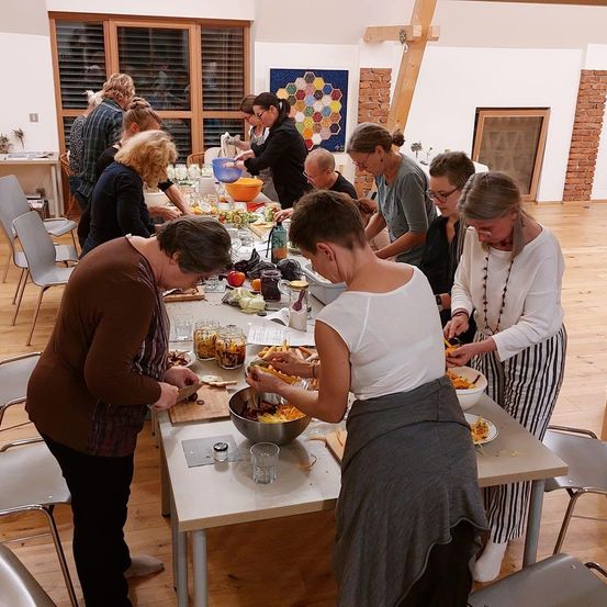 Eine Gruppe von Menschen versammelt sich um einen langen Tisch in einem Raum und bereitet Essen zu. Sie tragen Schürzen und scheinen an einer Kochaktivität beteiligt zu sein. Verschiedene Zutaten und Utensilien befinden sich auf dem Tisch.