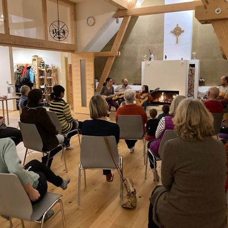 Eine Gruppe von Menschen sitzt in Stühlen um einen Kamin herum und hört zwei Personen zu, die Gitarre spielen. Der Raum hat Holzbalken und ein Kreuz an der Wand.