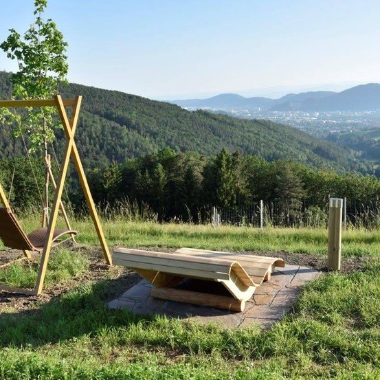 Eine Holzbank und Schaukel auf einem grasbewachsenen Hügel mit Blick auf Berge und eine Stadt in der Ferne.