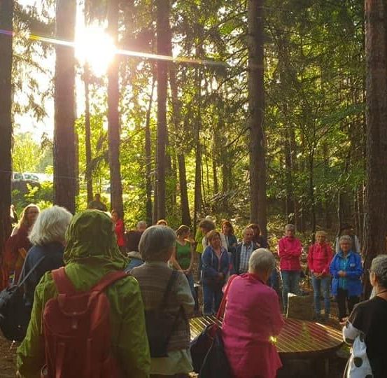 Eine Gruppe von Menschen in Jacken und mit Rucksäcken versammelt sich in einem Wald, wobei Sonnenlicht durch die Bäume filtert.