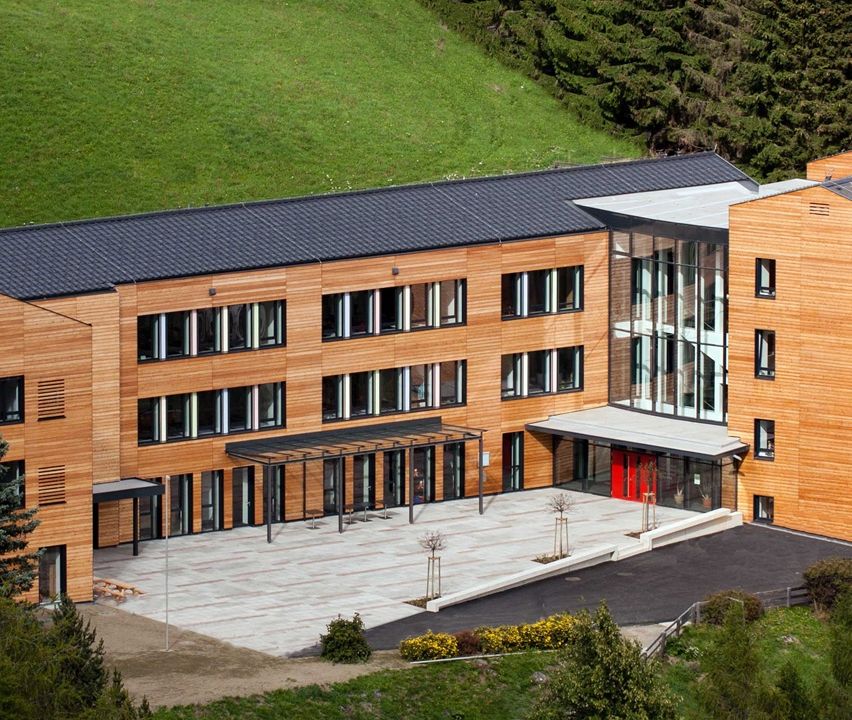 Eine Luftaufnahme eines modernen Bürogebäudes mit hölzerne Außenfassade, schwarzem Dach und vielen Fenstern. Es hat eine rote Tür am Eingang, einen gepflasterten Bereich davor und viel Grün.