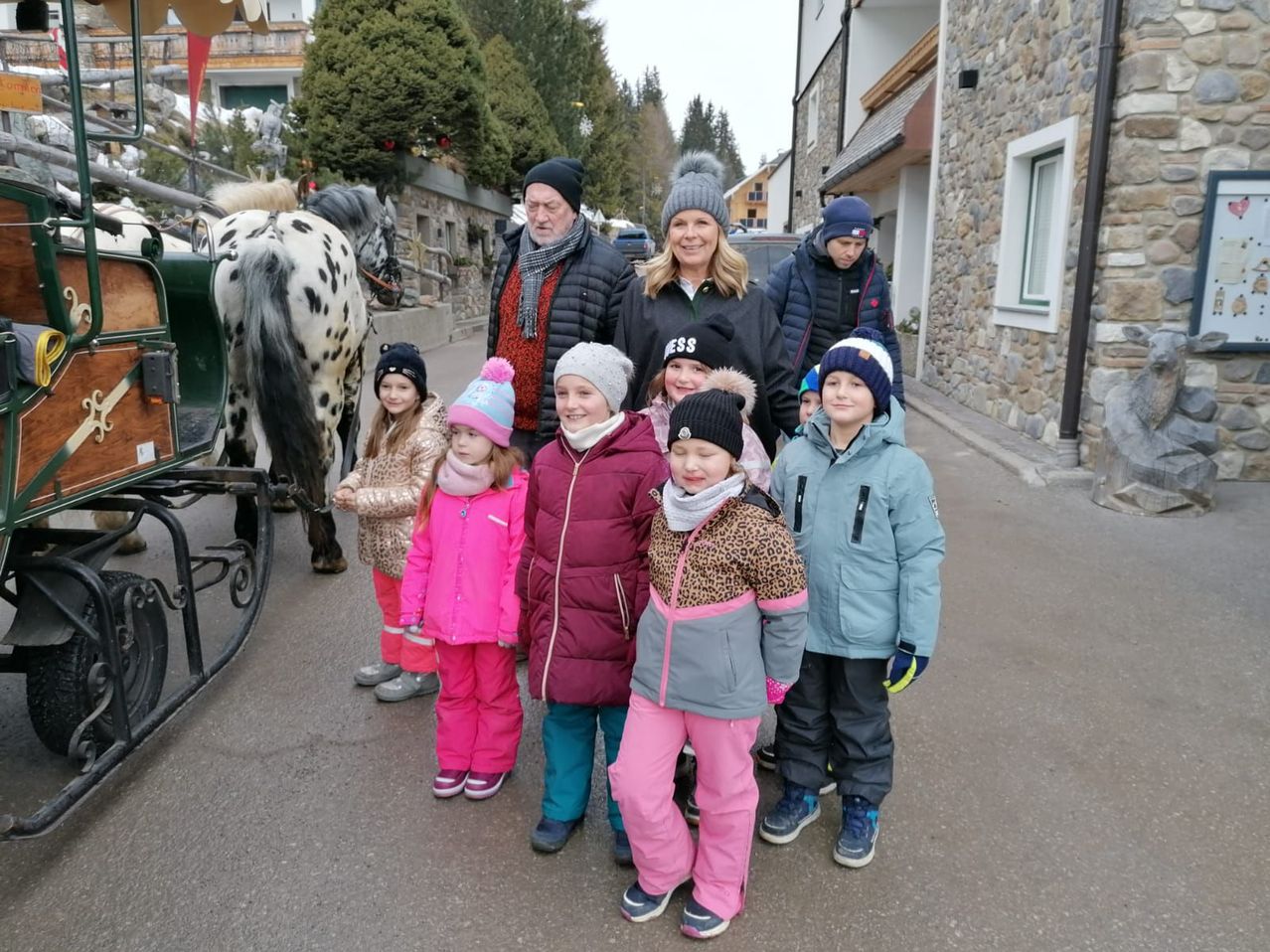 Eine Gruppe von Menschen in Winterkleidung, darunter Kinder, steht vor einer Pferdekutsche auf einer verschneiten Straße. Hinter ihnen ist ein Gebäude mit einer Steinmauer und einem Fenster zu sehen.