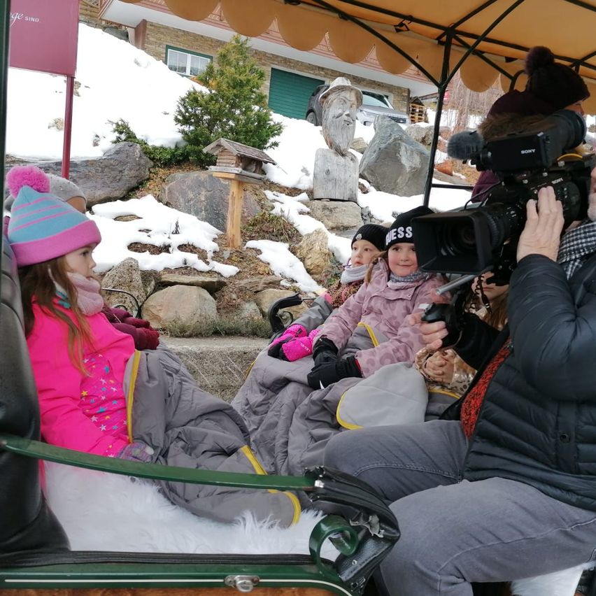 Drei Kinder in warmer Kleidung sitzen in einer Kutsche, die von einem Mann mit einer Kamera gefilmt wird. Die Kutsche ist von einem Baldachin bedeckt, und sie sind von Schnee umgeben.