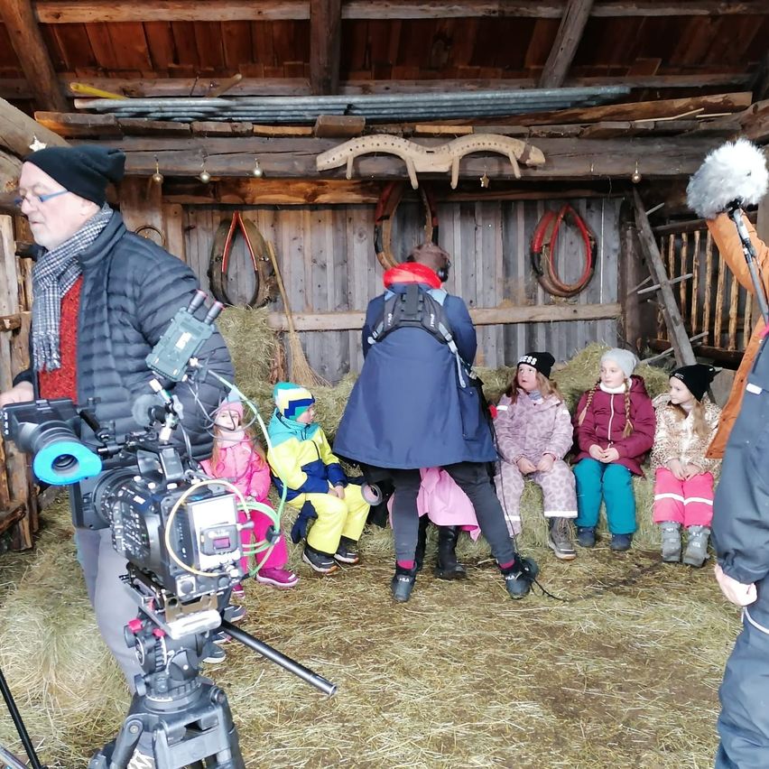 Eine Gruppe von Kindern sitzt auf Heu in einer Scheune, während ein Mann vor ihnen eine Kamera hält. Die Scheune hat Holzwände und eine Decke.