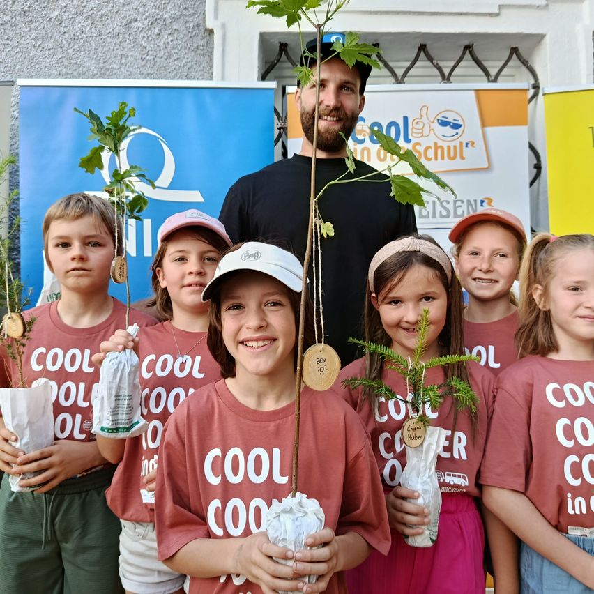 Eine Gruppe von Kindern und ein Mann halten kleine Pflanzen. Sie stehen vor einem Banner und einem Gebäude. Alle tragen rote T-Shirts mit dem Wort 'COOL' darauf.