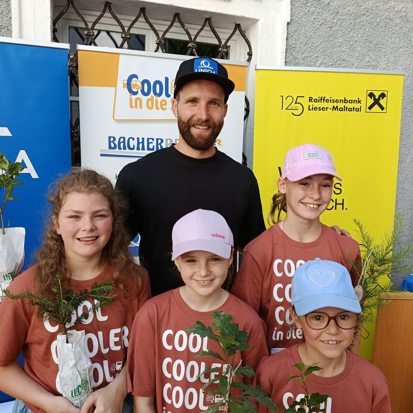 Eine Gruppe von Kindern und ein Mann posieren mit Pflanzen vor einem gelben Banner mit dem Text Raiffeisenbank Lieser-Maltal.
