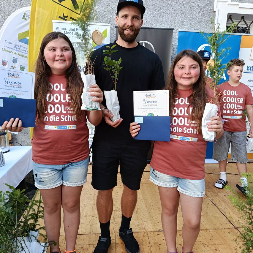 Zwei junge Mädchen und ein Mann stehen lächelnd zusammen mit kleinen Pflanzen in den Händen. Der Mann trägt ein schwarzes Shirt und eine Kappe, während die Mädchen burgunderfarbene Shirts mit weißen Shorts tragen. Sie halten Zertifikate in blauen Ordnern.