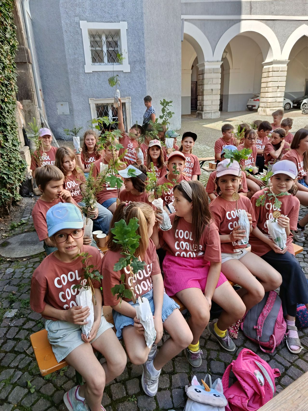 Kinder sitzen in einer Reihe und halten kleine Topfpflanzen in den Händen. Sie tragen passende T-Shirts mit der Aufschrift 'Cooler Cool'. Im Hintergrund befindet sich ein Gebäude mit einem geparkten Auto davor.